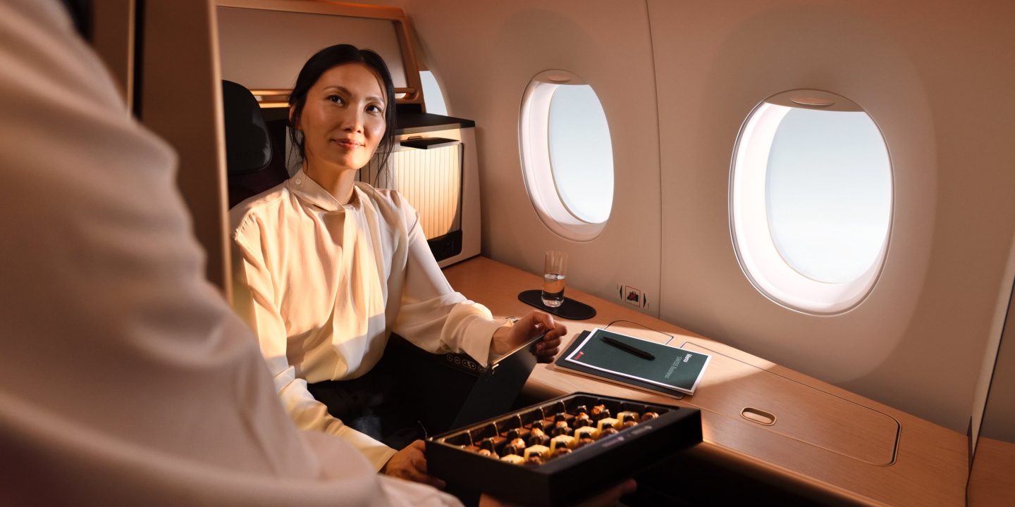 A smiling woman sitting comfortably in a premium airplane cabin seat next to two windows.